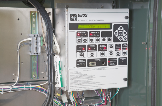 Una unidad de control de seccionamiento 6802 montada en la pared. La unidad se encuentra en la derecha de la foto, mientras que el lado izquierdo muestra los cables fijados a la pared que se conectan a la unidad. Los cables también sobresalen desde la base de la unidad y salen del área de la foto. La unidad de control de seccionamiento 6802 tiene un logo de S&C en la esquina superior izquierda de su panel frontal. El panel táctil frontal también tiene muchos botones etiquetados; "Interruptor 1" e "Interruptor 2" son legibles, los demás no. En algunos botones del panel táctil se iluminan luces rojas. 