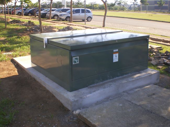 La unidad del interruptor Vista instalada por completo, con su caja verde oscuro con la marca S&C, se encuentra en su pedestal de concreto en primer plano. La evidencia del trabajo de instalación se encuentra presente, lo que incluye un palé de madera vacío en el fondo de la imagen, tierra recién removida alrededor del pedestal de concreto y materiales dispersos por el césped. En el fondo se observa una línea de árboles, una fila de vehículos estacionados y una calle.