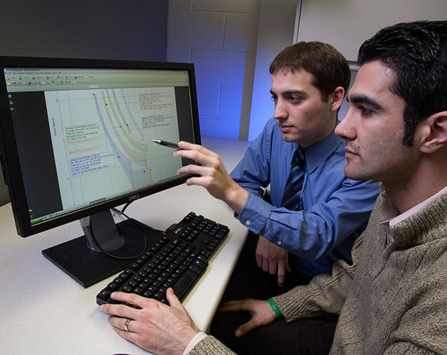 a man pointing at a computer screen with a pen wearing a blue shirt and dark tie to a man looking at a tcc curve on the screen wearing a white collar shirt and tan sweater.